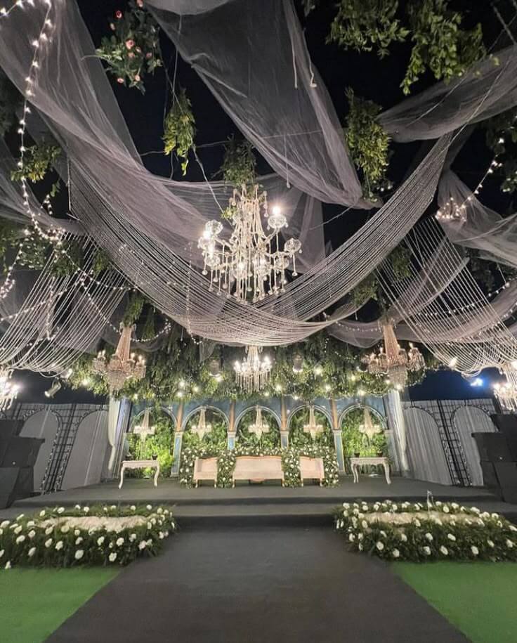 Luxury wedding stage setup with floral backdrop, hanging chandeliers, draped ceiling decor and elegant seating arrangement by professional event planner.